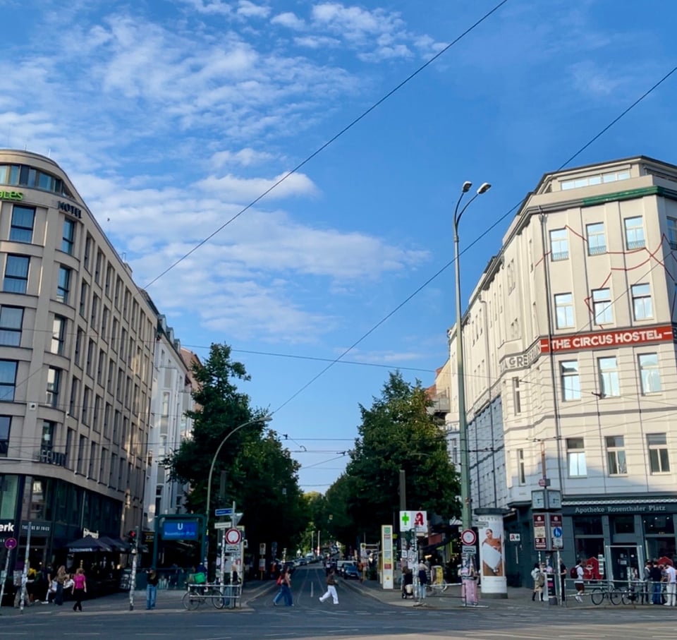 Shows an intersection at one of the busy districts in Berlin. On each side is a 5 story building on the edge of the street in focus. It is late afternoon and the building on the right is illuminated with bright setting sun. The street is lined with lush trees. There are people crossing the road while the pedestrian crossing light is on.
