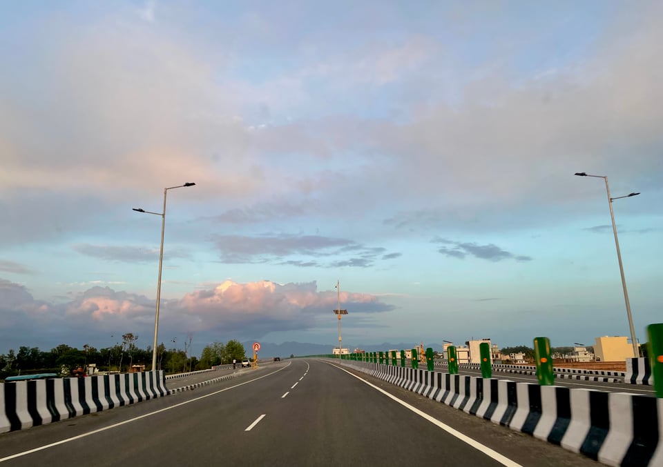 Shows a highway slightly wounding towards the left at the far end that the photo captures. There are red and white evening clouds in the backdrop of a pale blue and grayish sky. Two street0lights flank both sides. There is zebra coloured pattern on the stone dividers on both sides of the road
