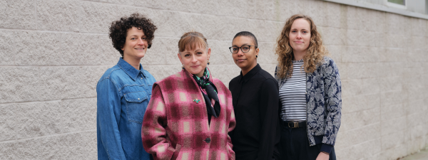 Four women standing and facing the camera. From left to right is a woman with curly hair in a denim shirt. Next is a slightly shorter woman, with a pink and white coat, and a full fringe. Next is a woman in a black shirt, black glasses and a buzzcut head. Finally on the far right is a taller woman in a blue and white jacket, with a stripy top underneath. She has curly brunette hair.