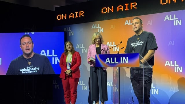 Three people stand on stage at a conference for an announcement. A man is at the podium, and two women stand beside him.