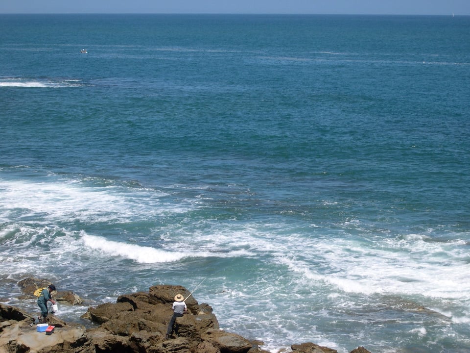 On the lower left hand side of the frame we can see some rocks. One person is sorting out the fish they have caught. While the other is handling the fishing rod out into the ocean. The ocean is relatively calm but the waves break near the rocks.