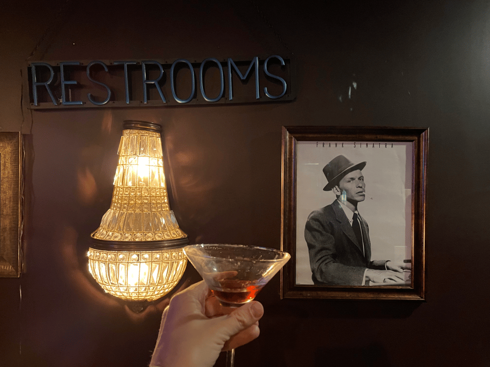 A cocktail glass in front of a picture of Frank Sinatra and a restroom sign