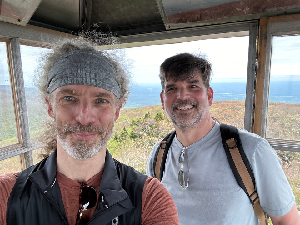 Photo of two white men standing in a room at the top of a fire tower, with woods and mountains behind them