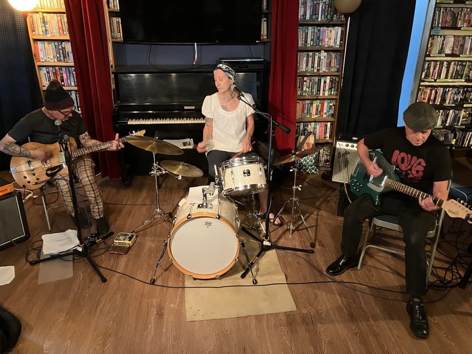 A woman in a white t-shirt sits behind a drum kit, flanked on either side by men in black shirts playing electric guitars.
