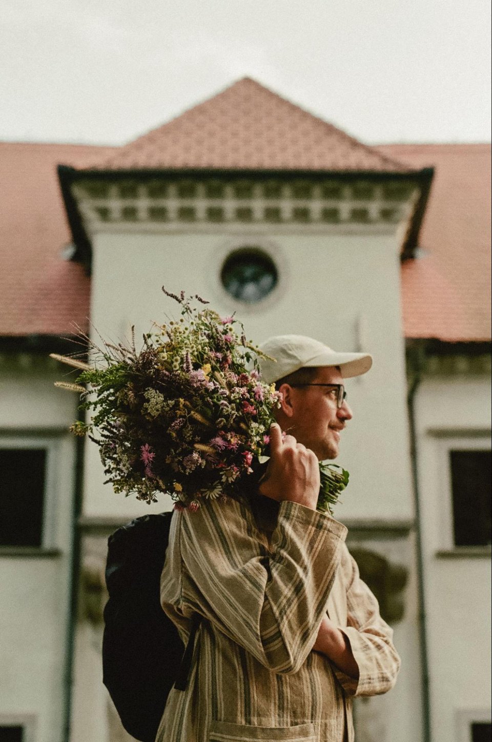 [Kristoffer wears a white hat and a tan striped shirt, holding a full bouquet of wildflowers in front of the white walls and terracotta roof of the castle.]