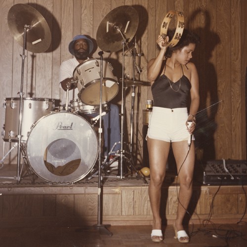 A young Black woman wearing a black camisole and white shorts, holding a microphone and shaking a tambourine. She's standing in front of a drum kit being played by a Black man wearing a hat