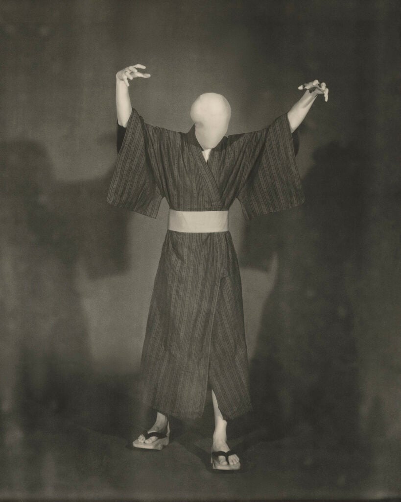 A sepia-toned vintage photograph shows a figure in a striped kimono with a pale sash, wearing wooden sandals, standing with both arms raised and fingers splayed; the face appears as a blank, featureless white mask, and soft shadows fall on the mottled backdrop behind.