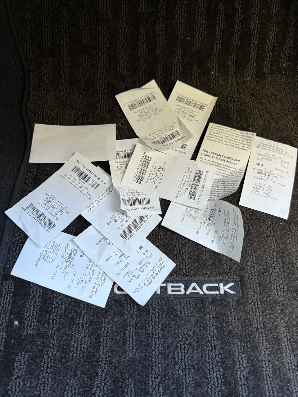 scattered receipts and tickets from hospital parking garages, on the black floor mat of a Subaru Outback