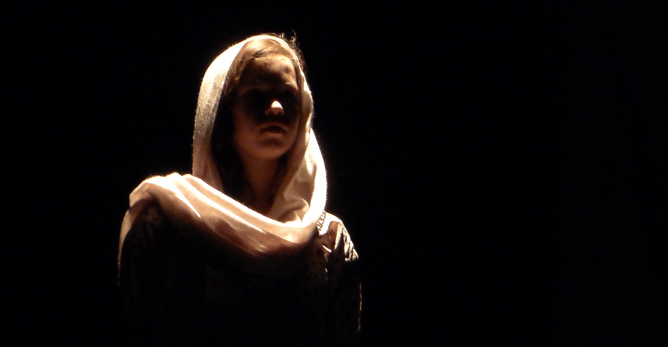 A figure wearing a light-colored headscarf stands against a dark background, illuminated from above.