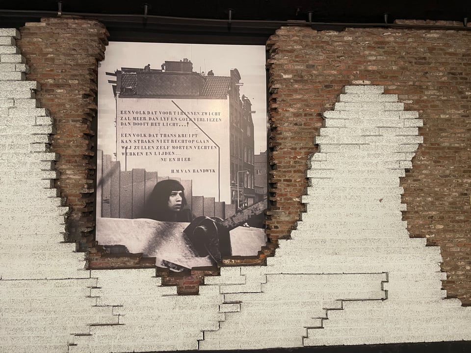 An exposed brick wall in the metro of Amsterdam has a large photograph on display. The photograph is of the side of building with a Dutch poem painted on the white wall.