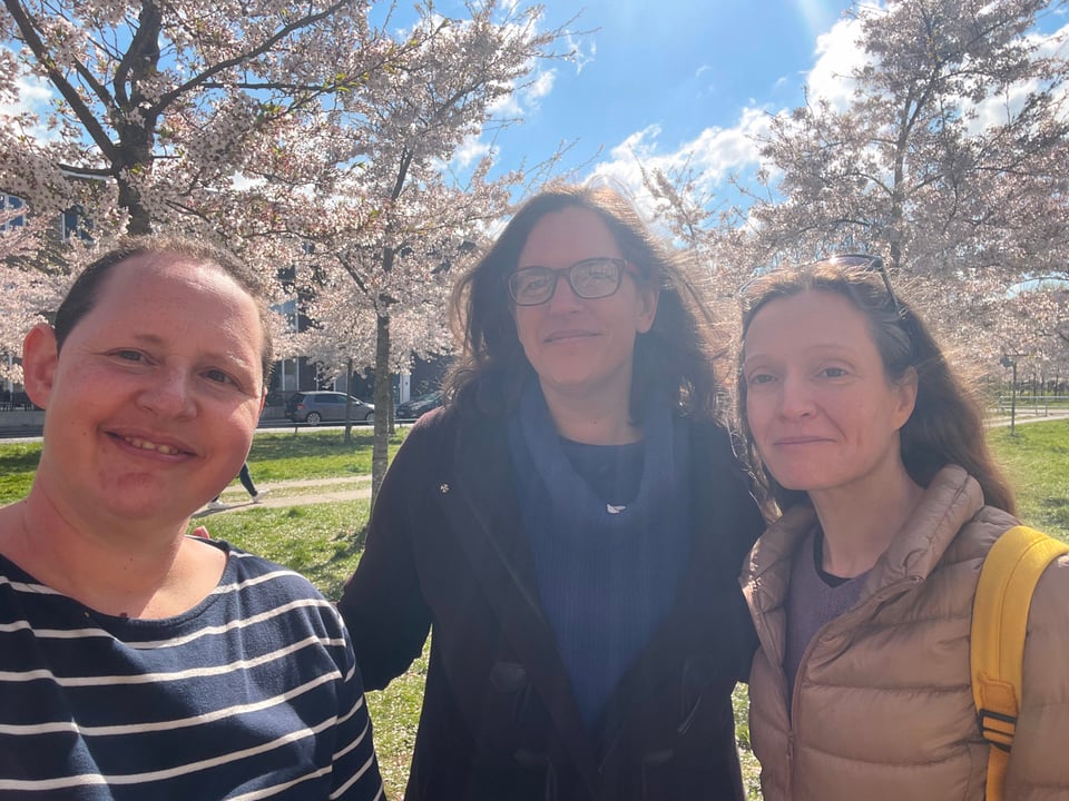 Three women look at the camera where cherry trees in full bloom fill the background behind them.