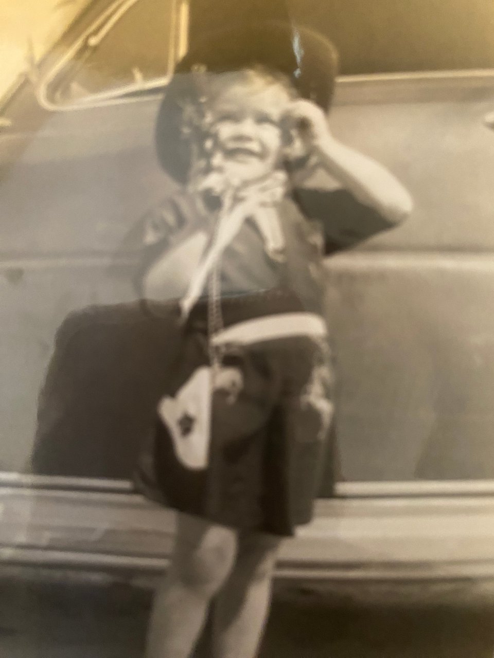 Black and white photo of a little girl dressed in a cowgirl outfit in front of a car