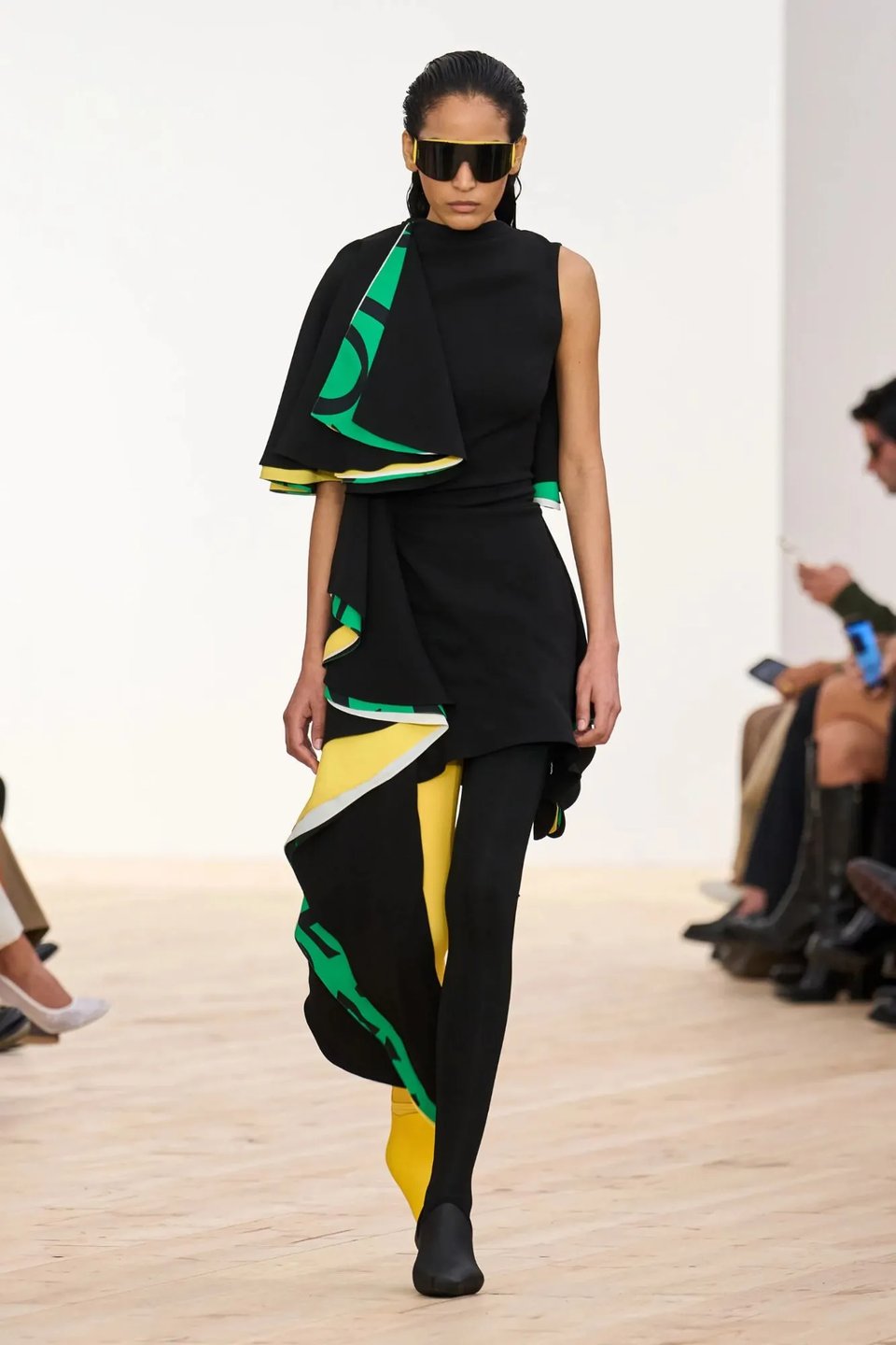 Model in a fabulous Loewe suit, designed with asymmetrical ruffles and color pattern of black/green/yellow design, including one black pant leg and one bright yellow pant leg. The model also wears big matching black sunglasses with yellow rims. The whole look is very futuristic. Very Tron or Fifth Element.