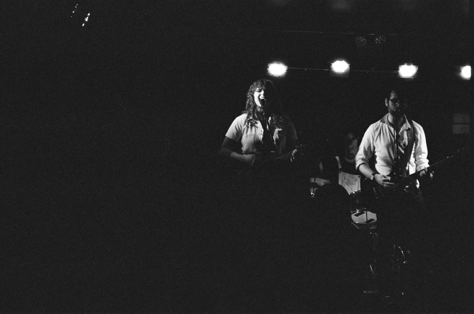 A black and white film photo of the band Sorry Darling. There's a large empty space of darkness on the left. You can see Liz + Steve singing and playing guitar on the right, and Dan behind them playing drums.