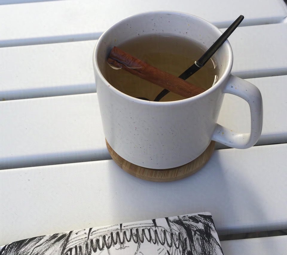 A cup of tea in a white ceramic cup on an outside white table. There's also a corner of a sketchbook visible in the frame.