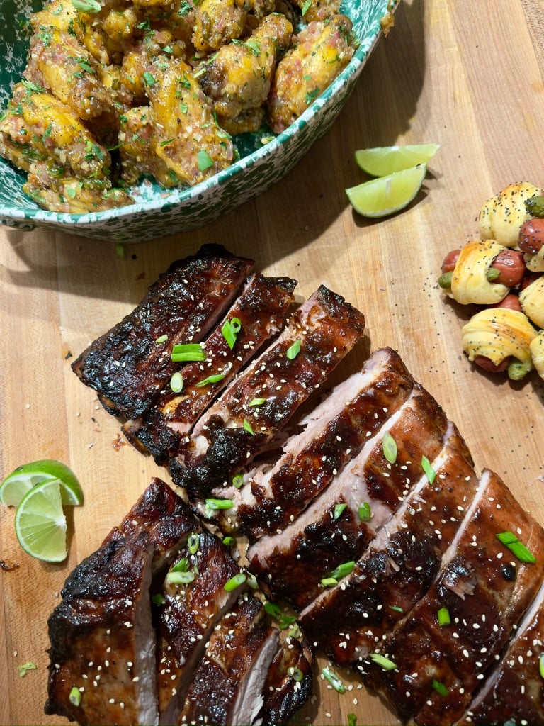 A rack of lacquered ribs in the foreground flecked with sesame seeds and scallions with a platter of lemongrass wings and small pigs-in-a-blanket accompanying.