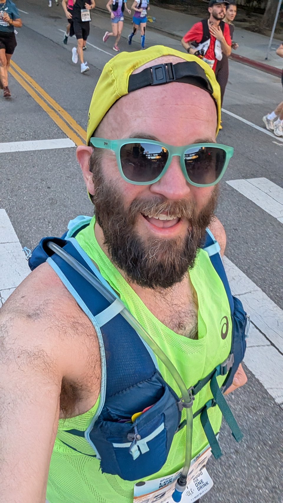Classic mid-race selfie. I'm in a yellow Tribe hat, greenish Goodr sunglasses, hydration vest, and a neon green singlet.