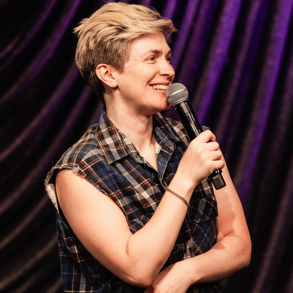 Nora smiles fondly out at the audience while speaking into a mic and wearing a very butch plaid button-up with the sleeves cut off.