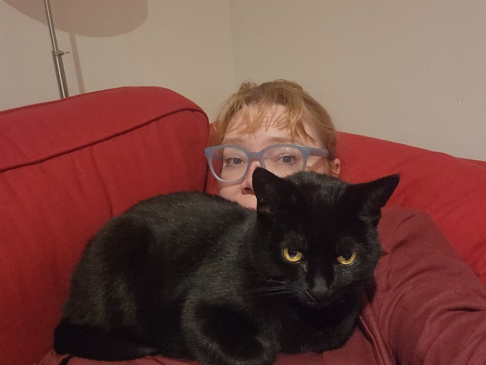 Kate looking tired in a red linen jacket, reclining on a red couch. 10 pounds of black cat is on her chest.