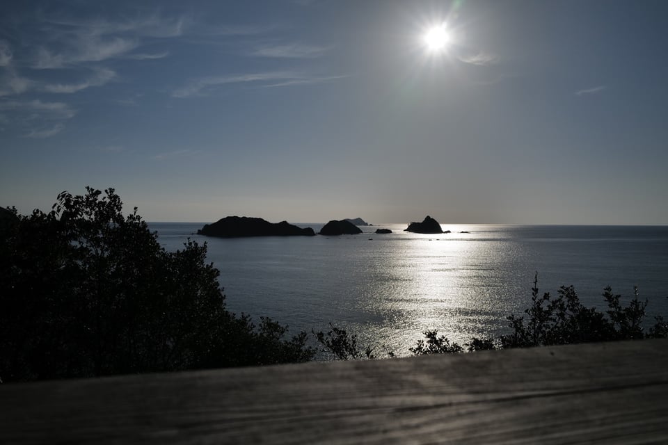 A seascape photograph taken from an elevated vantage point showing silhouetted rocky islands in calm waters under a bright sun. The sun's reflection creates a shimmering path across the sea's surface. Vegetation and shrubs frame the foreground on both sides, appearing dark against the luminous water and sky. The sky is predominantly blue with some wispy clouds. A wooden railing or deck edge is visible in the immediate foreground.
