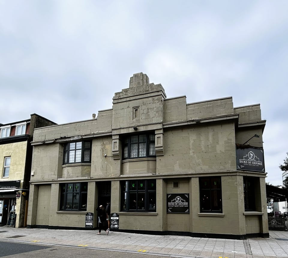 A muddy grey/brown pub. It's two storeys high. It has a heavy stepped pediment and the straight lines of a Deco building but then it also has inset bay windows with mullions like it's an Arts and Craft house.