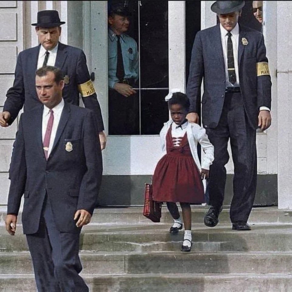 A small Black six-year-old girl wearing a red dress, white cardigans, and dress shoes is escorted down stone steps by three men in suits wearing yellow armbands that read "U.S. MARSHAL."
