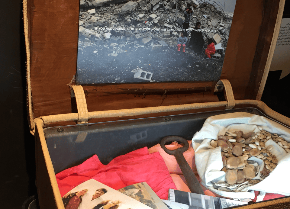 Photograph of an open vintage suitcase containing family photographs, papers, red fabric, a large iron key, and a cloth pouch filled with coins. Lid-mounted screen shows rubble, and a family of 4 sitting in the rubble, and a prompt asking IF YOU HAD 30 MINUTES BEFORE YOUR HOME WAS DEMOLISHED, WHAT WOULD YOU SAVE?