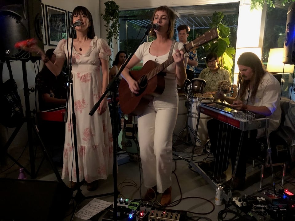 Photo of Swimming Bell playing live at Oblivion. There are seven musicians squeezed onto a makeshift stage at the front of a store. Two singers are at the microphones. In the background are a musicians playing pedal steel, drums, conga and other instruments.