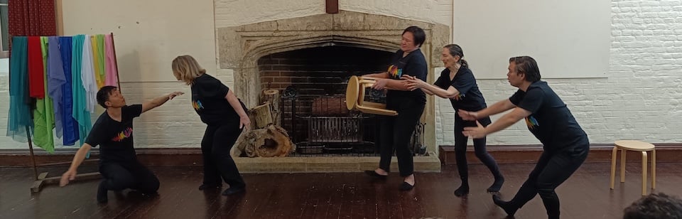 A group of five performers in matching black t-shirts strike dramatic poses in a hall with a large stone fireplace, with colourful scarves draped over a rack on the left, a wooden stool on the right, and one performer holding a small wooden stool in the centre.