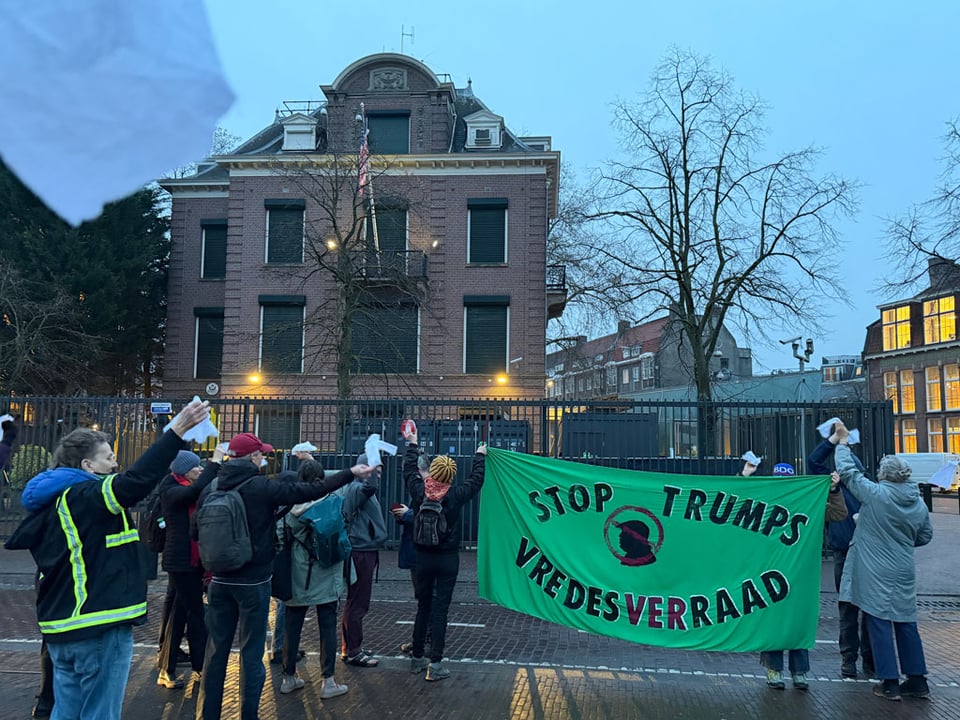 A group of people waving white handkerchiefs stand in front of the USA Consulate in Amsterdam. Two hold a large green sheet that says "Stop Trumps Vredesverraad" (Stop Trump's Board of Peace").