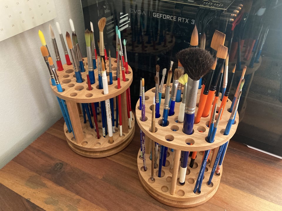Two wooden paintbrush organizers on a brown, wooden desk next to a computer. The paintbrush organizers are both round with several spaces for brushes, of which there are many in various sizes and styles in each organizer.