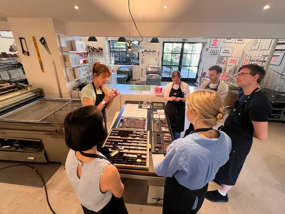 Photo of a group of people standing around a desk, looking at metal type samples, intently listening to one person explaining