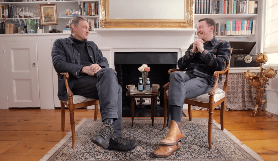 Screenshot from a video of two men sitting in chairs and facing each other. There are bookshelves in the background.