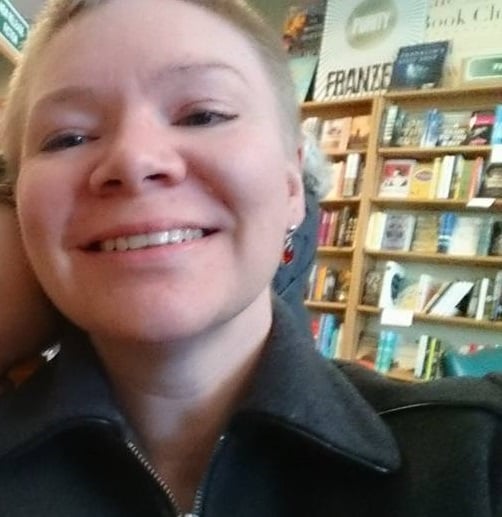 Kate with very short hair, grinning, slightly out of focus, bookshelves behind.