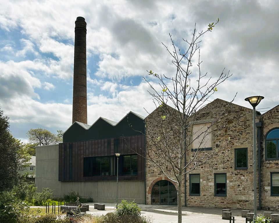 A factory building with a tall brick chimney. The building has the classic zigzag roof. The right side is in stone, with an arched doorway and somewhat random windows. The left side has been built (or rebuilt) to match the roof line but is a concrete block with copper clad walls and regular windows.