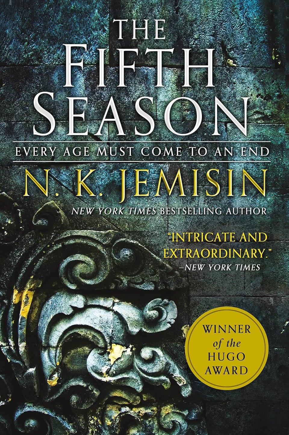 Cover of The Fifth Season by N.K. Jemisin with additional text subtitling "every age must come to an end," noting that Jemisin is a New York Times bestselling author, providing the blurb "intricate and extraordinary" from the New York Times, and adding that this book is a winner of the Hugo award, all over the background of a pitted and viridescent stone wall over an ochre-rusted gray metal sculpture