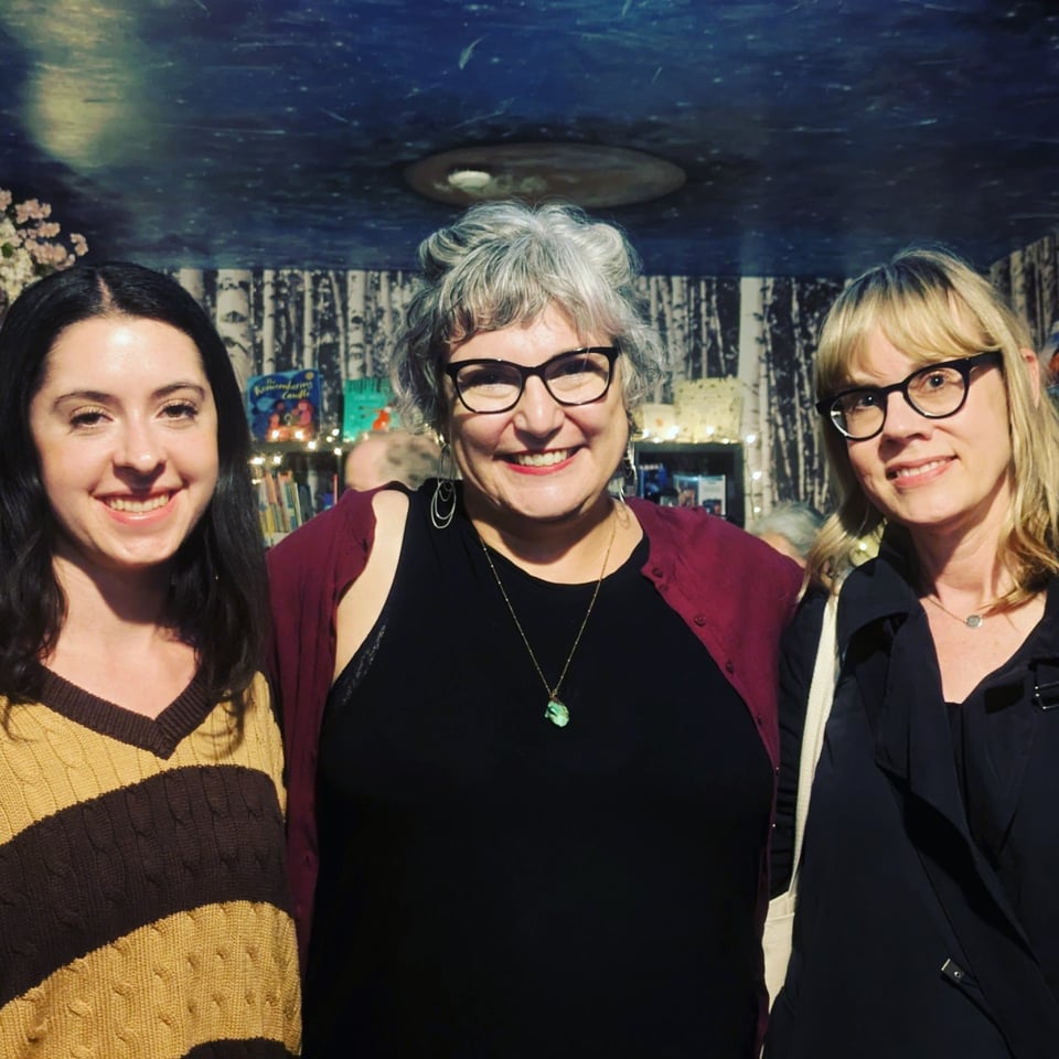 Three white women stand together after the poetry reading. They are smiling and standing in front of a bookshelf.