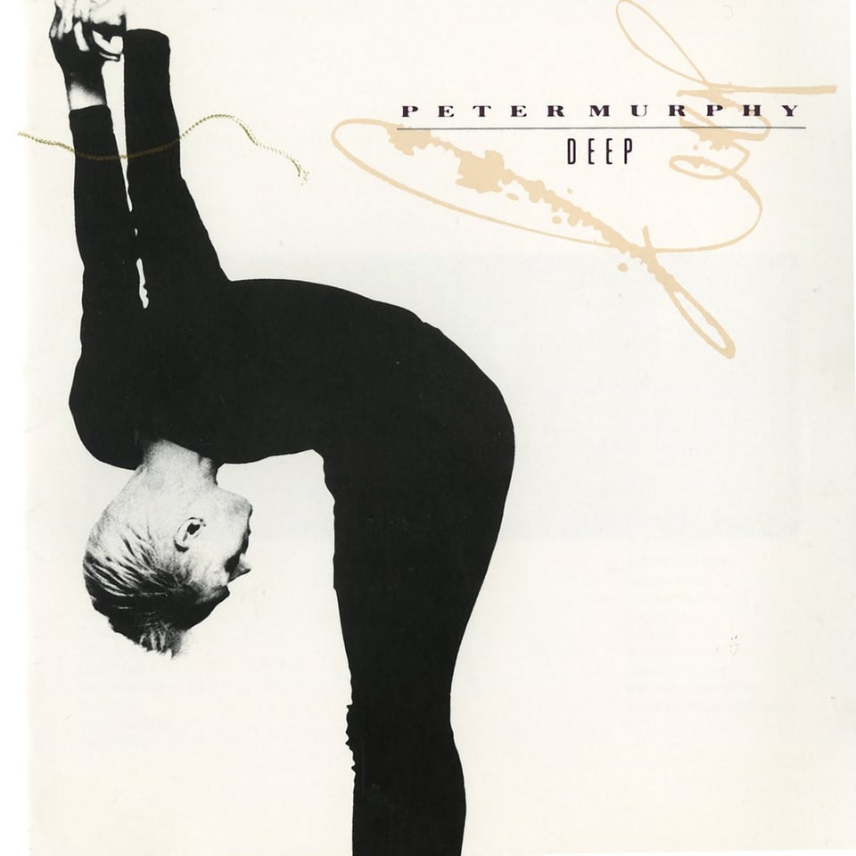 A black and white photo of Peter Murphy, a white man with short blond hair in a black leotard, bending at the waist until his head is upside down and facing his crotch. His arms are extended straight up behind him, hands clasped, on the same vertical plane as his head. This is on a blank white background.