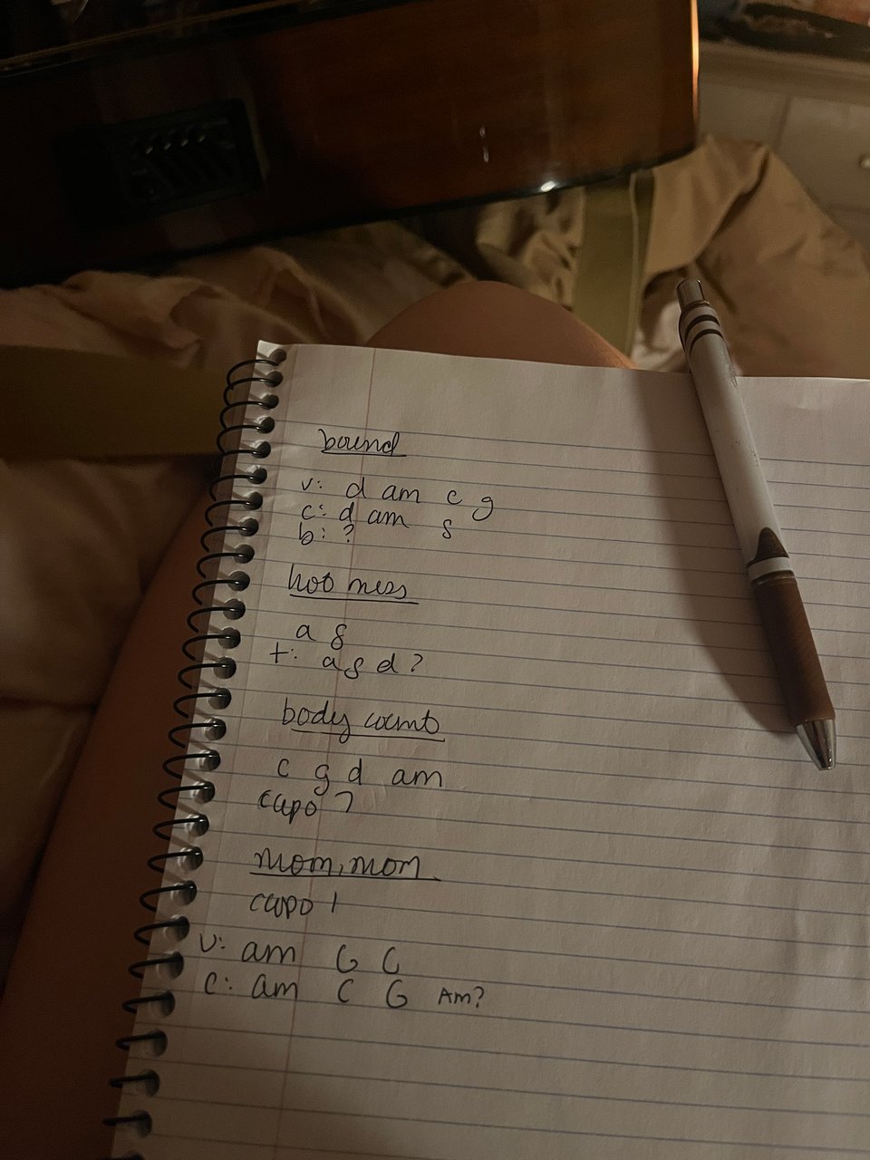 notebook page with song names (bound; hot mess; body count; and moon, moon) and tentative chords in low light on Nola's lap, guitar in the edge of the shot