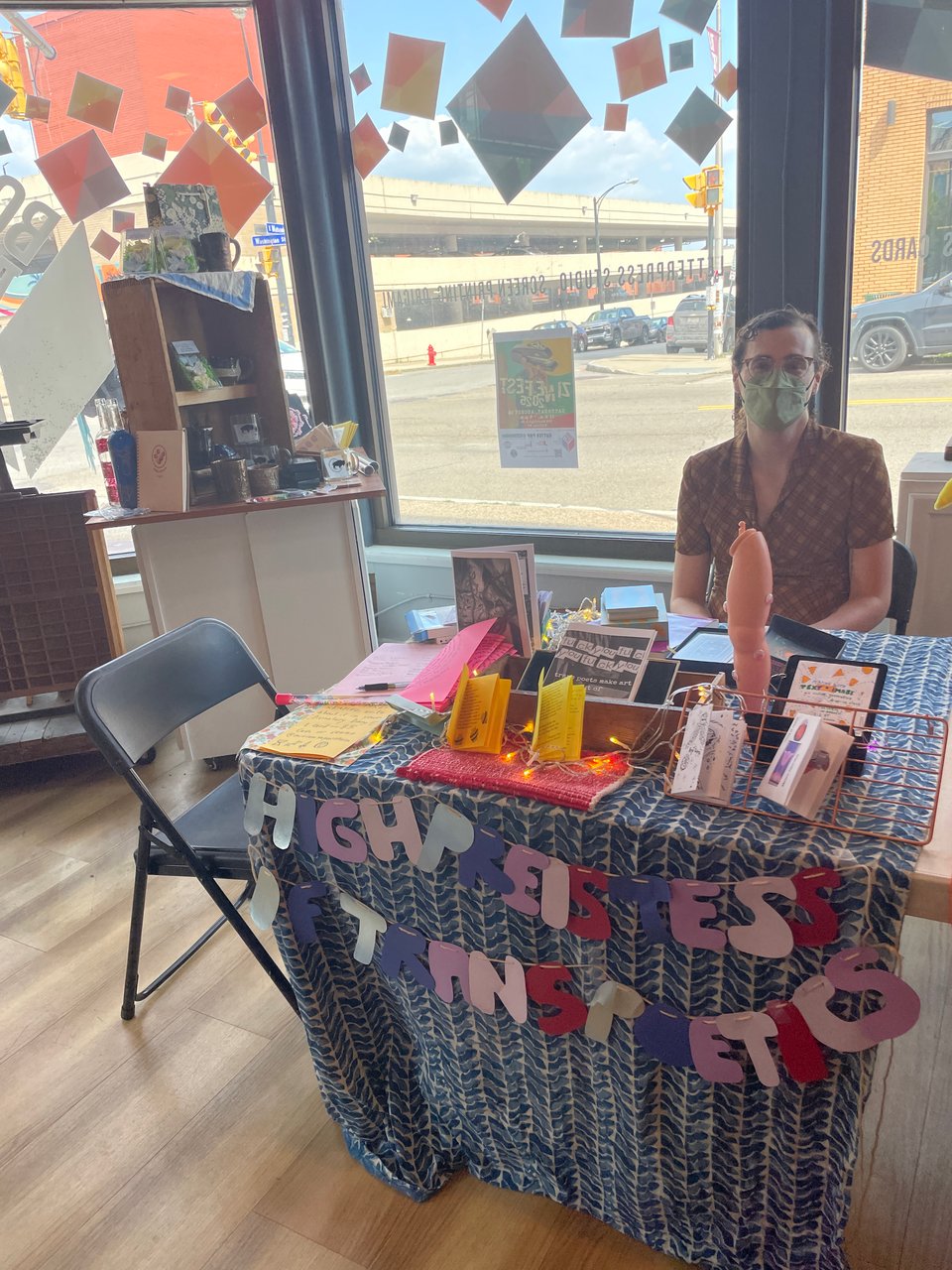Zoe, a masked white woman in a brown dress, sits at our zine table which has a blue tablecloth and is covered in paper zines, with a banner that says “high priestess of trans poetics” in front of a decorated store window