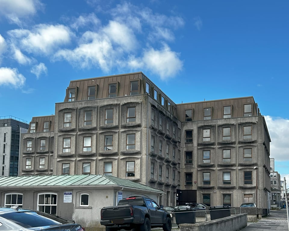 The back of the same building, showing is is in the form of a T. There's a lovely blue sky but overall the building is drab. Not helped by the carpark and a low concrete building in the foreground.