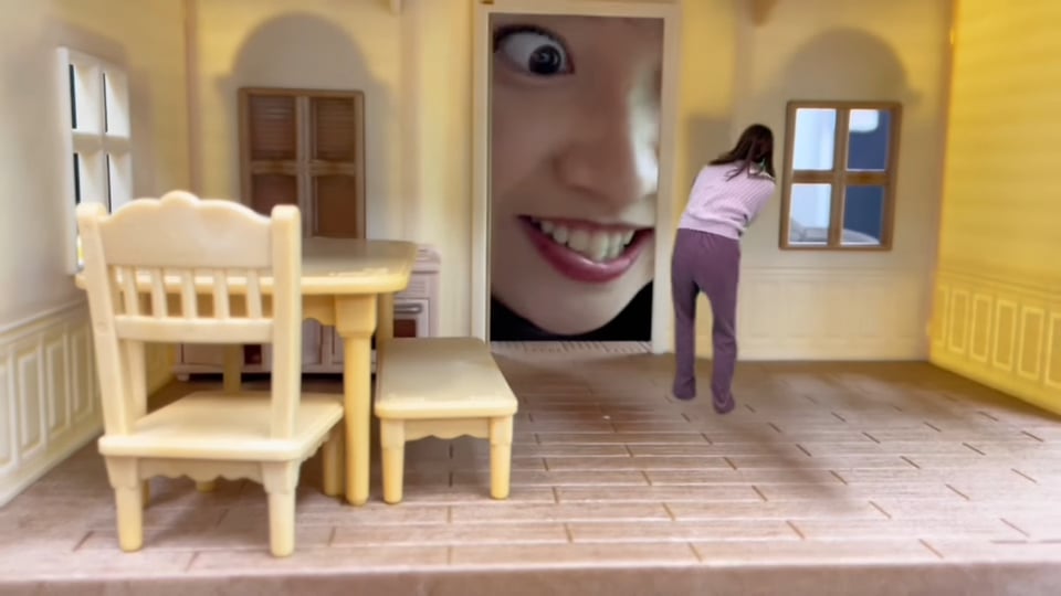 A tiny woman cowers in the living room of a plastic toy dollhouse while her friend's crazed expression fills the doorway