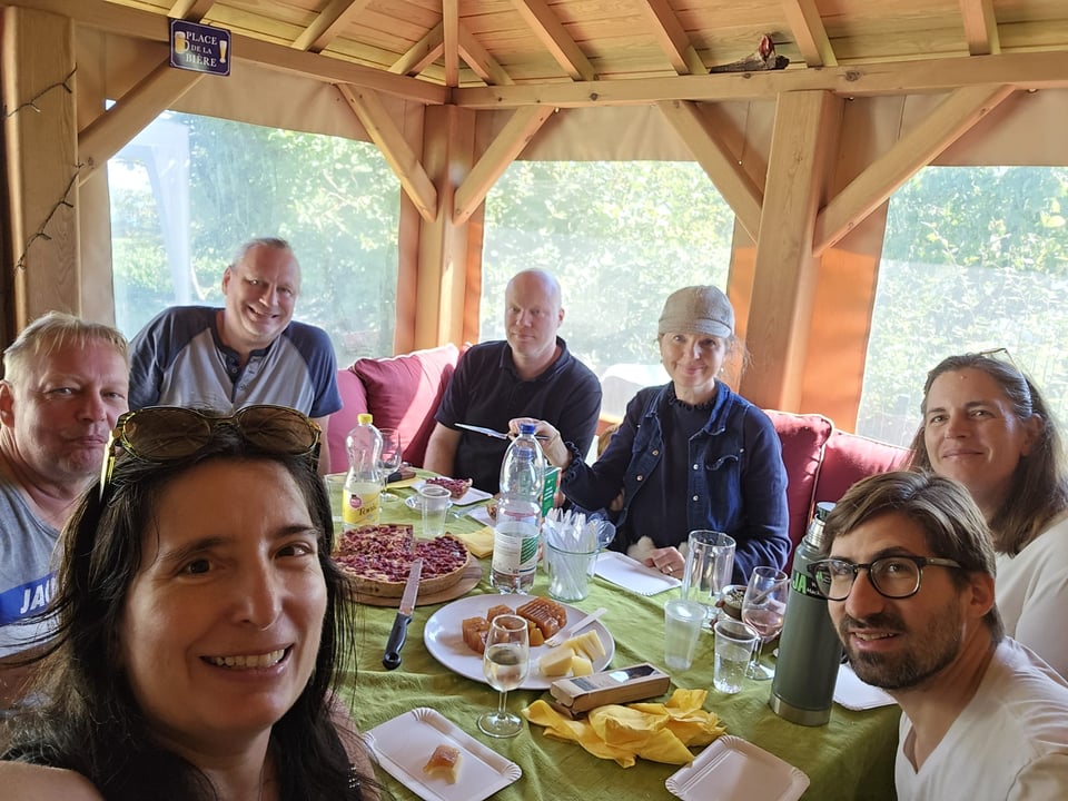 Seven people sit around a potluck table inside a wooden cabin with large light-filled windows