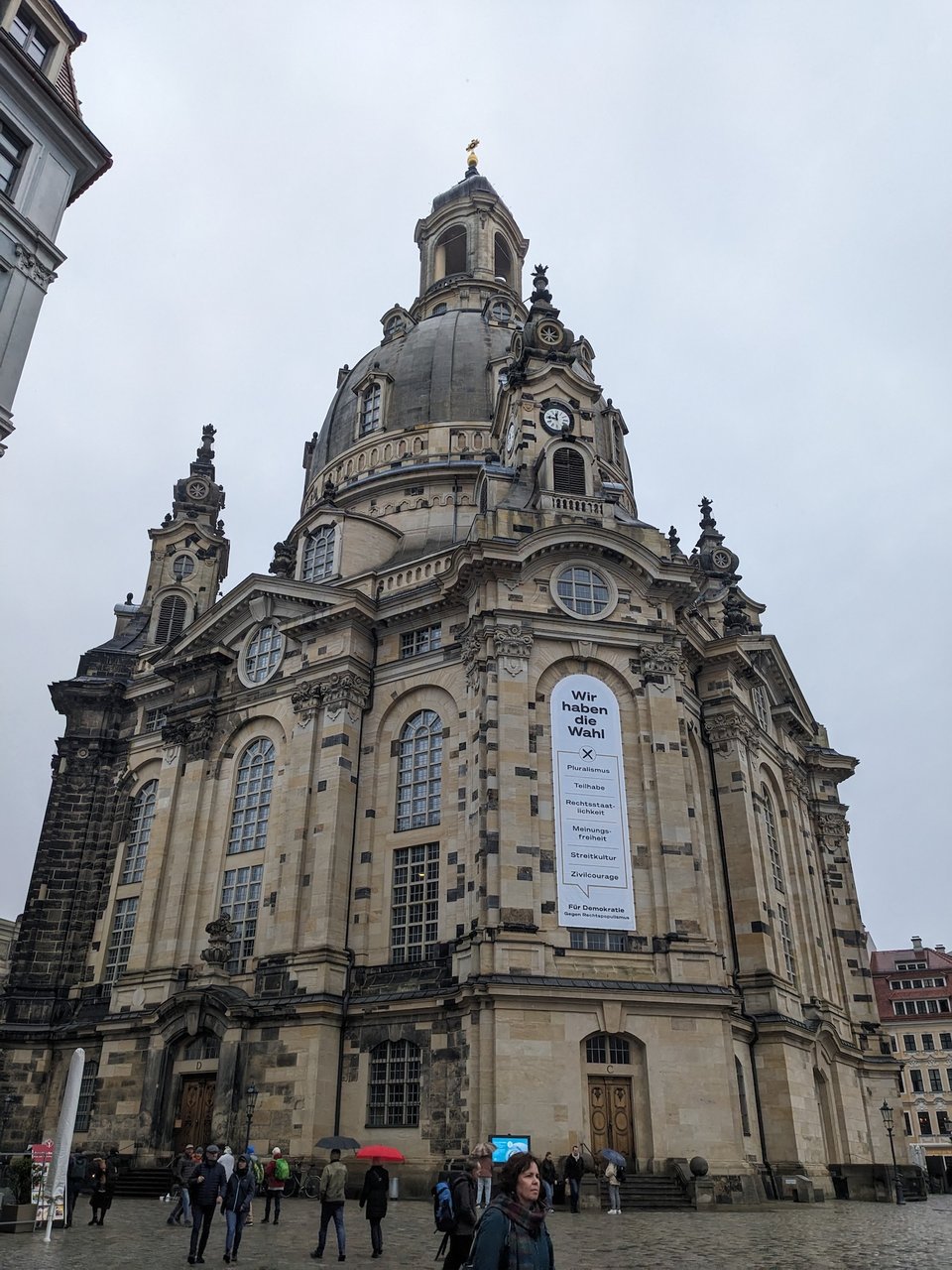 A picture of the Dresden Frauenkirche. It is a roughly hexagonal sandstone building with a bell-shaped dome. The support pillar on the left is black with soot. Most of the bricks are tan, but some are black. The black ones are from the original structure. They represent less than 25% of the visible bricks. The sky is solid grey.