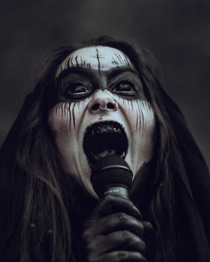 Photo of a black-haired woman singing/screaming into a microphone. The hand holding the microphone is painted black, as is her neck and the area around her eyes. She has a small septum piercing and her eyes and teeth are black.
