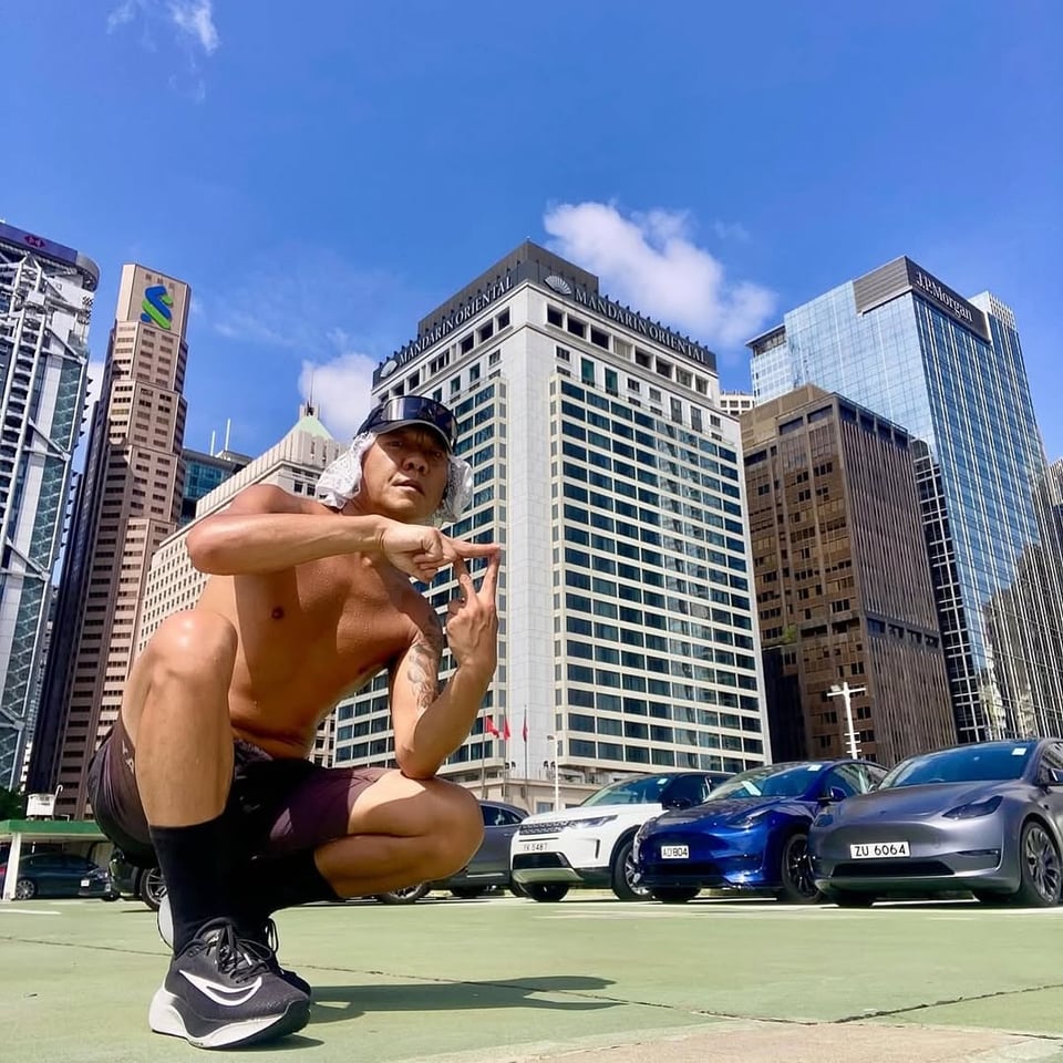 Calvin Yu, shirtless, in a baseball cap and purple shorts crouches in front of a downtown cityscape, making a hand sign. Behind him are tall office buildings including the InterContinental hotel, luxury cars, and a bright blue sky with white clouds. The photo is taken from a low angle a rooftop.