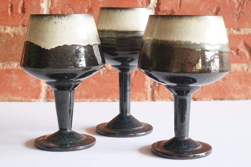 Three large wine goblets with stepped bases and slightly flared stems up to round-bottom bowls that angle in from a wide base to a narrower rim. glazed in a deep blue black from the base, turning white in the top half of the cup