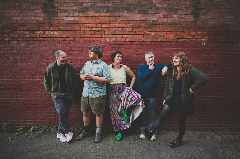 Three men and two women stand in front of a brick wall. Each woman has one foot off the ground, as if they're comparing footwear.