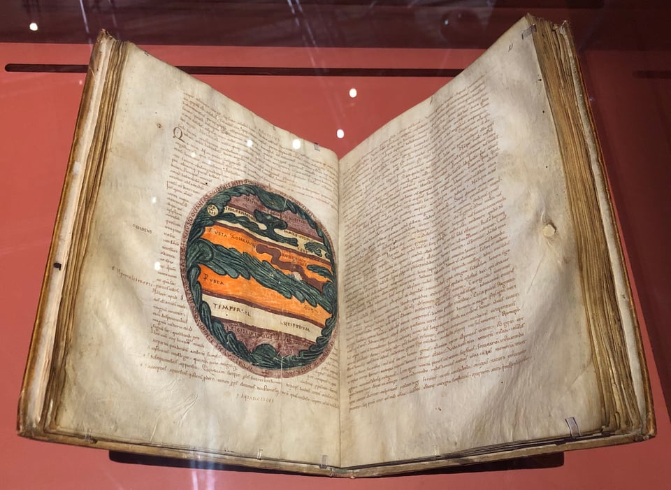 A medieval manuscript in a museum case. Halfway down the left-hand page is a large, circular diagram of the world. It’s divided into five curly, horizontal zones. The far northern and southern zones are a purplish colour. The next zone in is white. The central zone is a bright orange, and bisected by swirling, blue-green waters which also surround the globe’s rim.
