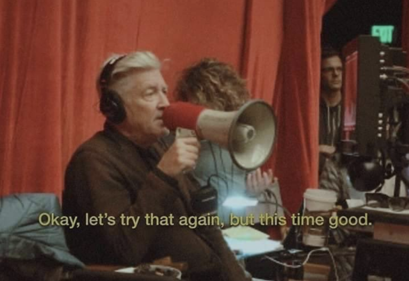 A still shot of David Lynch with a megaphone on set with the words "Okay. let's try that again, but this time good."
